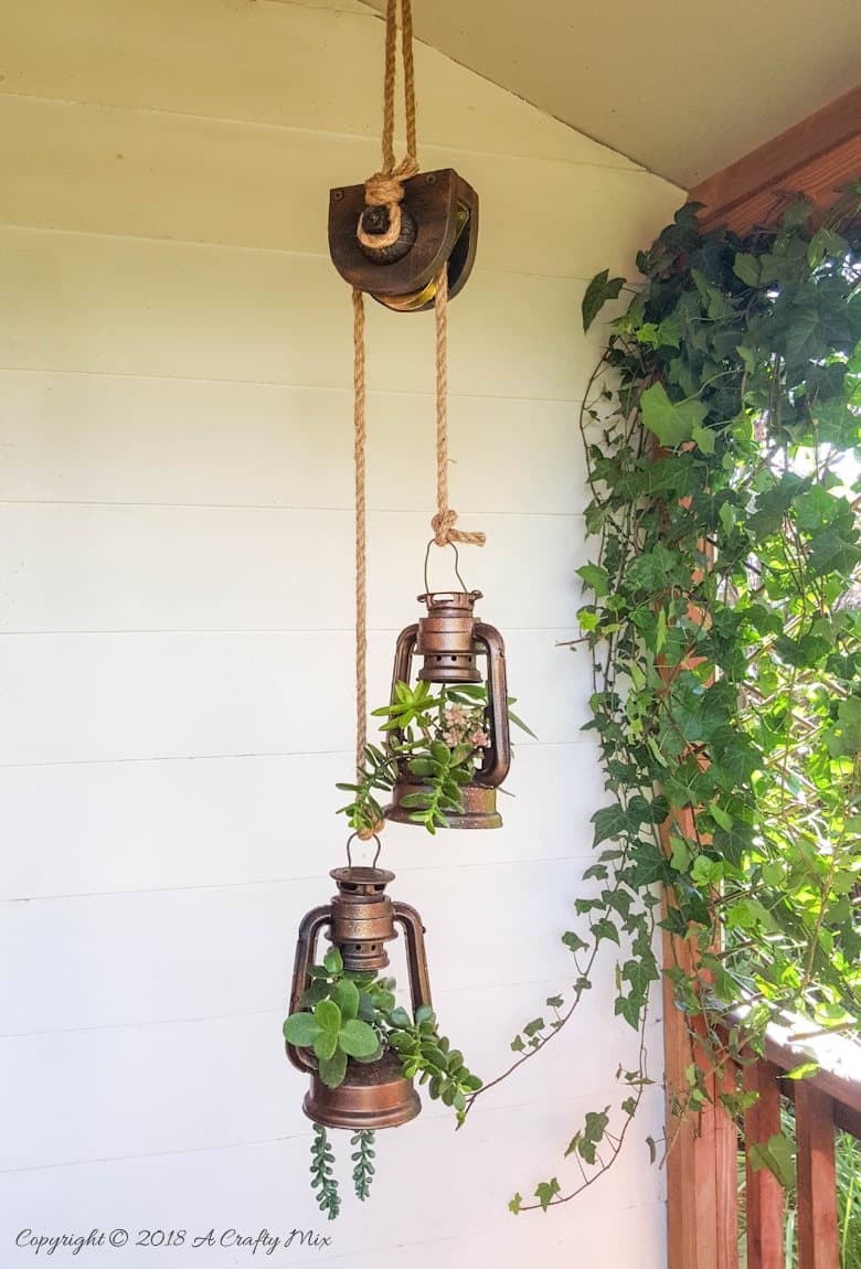 DIY lantern planters hanging from a faux antique pulley on a farmhouse porch. 