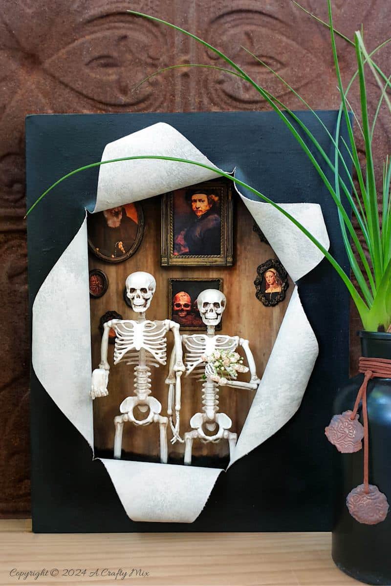 Get your spook on and add a touch of macabre whimsy to your décor with this DIY Halloween RIPped canvas tutorial. It even lights up. We’ll show you how to turn a deep canvas into an eerie masterpiece using paint, paste and two loving skeletons. Oh, be warned; we will be cutting, slicing, and mangling things up a little to make this haunting diorama.