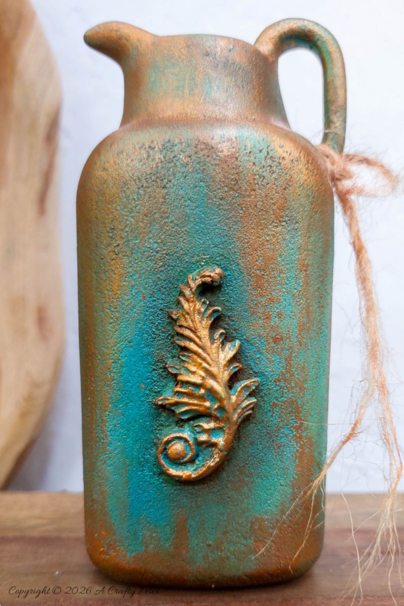 Vertical image of a faux verdigris patina vase made from an upcycled baby powder bottle, with dried grasses arranged in the jug‑style vase,