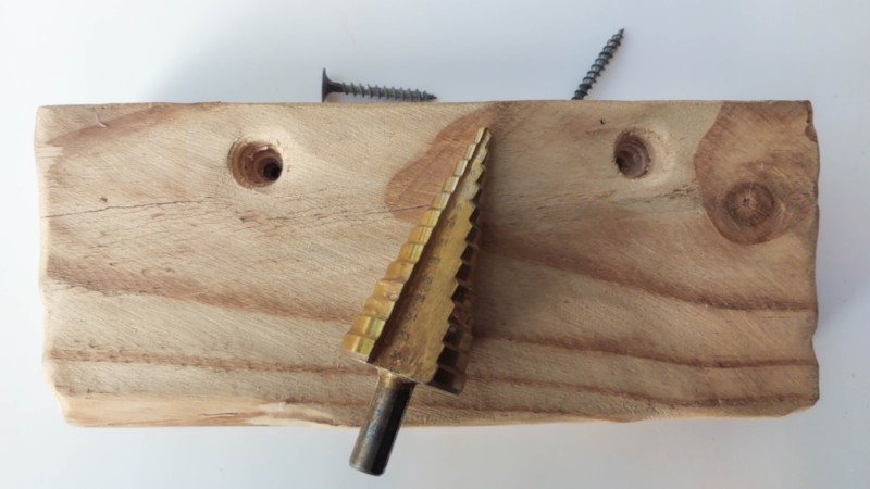 A step bit lying on top of a piece of wood. There are two countersunk holes in the wood and wood screws in the background.