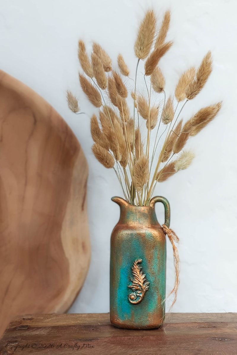 Vertical image of a faux verdigris patina vase made from an upcycled baby powder bottle, with dried grasses arranged in the jug‑style vase,