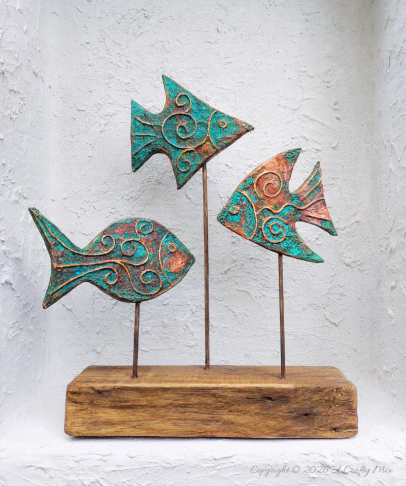 Three rustic patina fish on wooden sticks used as DIY coastal decor, crafted from layered cardboard and copper tape.