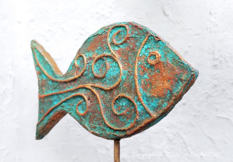 A rustic patina fish on a wooden stick used as DIY coastal fish decor, crafted from layered cardboard and copper tape.
