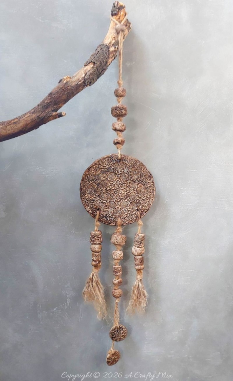 Rustic clay medallion wall hanging tied to a weathered tree branch with jute twine, featuring a strand of textured clay beads above the embossed round medallion against a soft gray background.