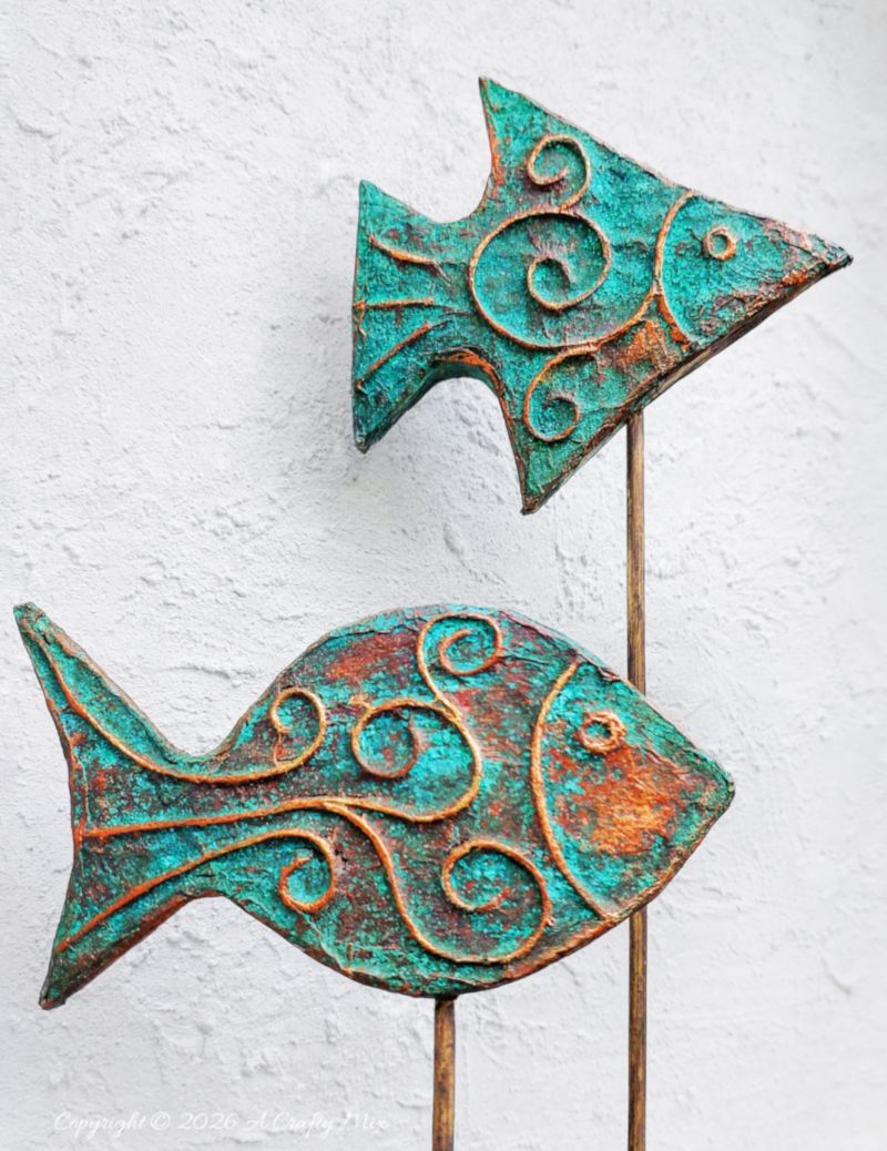 Two rustic patina fish on wooden sticks used as DIY coastal decor, crafted from layered cardboard and copper tape.