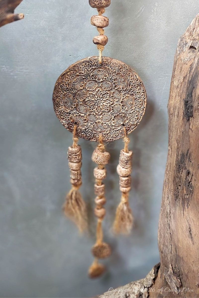 Rustic clay medallion wall hanging tied to a weathered tree branch with jute twine, featuring a strand of textured clay beads above the embossed round medallion against a soft gray background.
