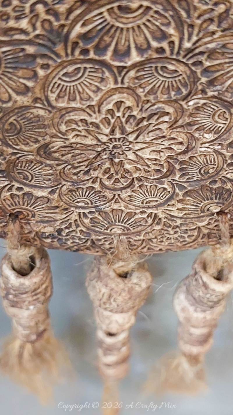 Close‑up of a rustic clay medallion wall hanging showing an intricate mandala pattern with darkened recesses and three jute‑wrapped tassel strands dangling from the bottom.