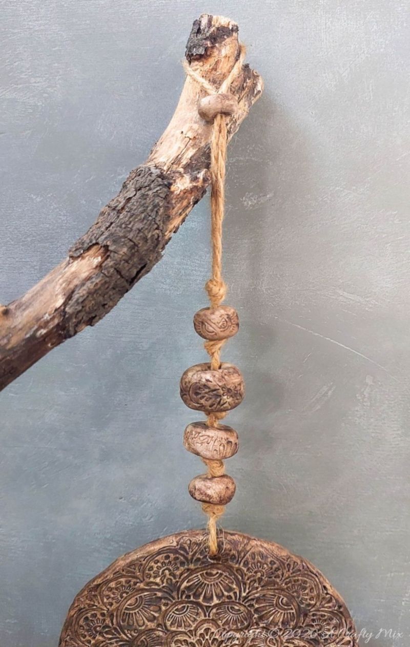 Rustic clay medallion wall hanging tied to a weathered tree branch with jute twine, featuring a strand of textured clay beads above the embossed round medallion against a soft gray background.