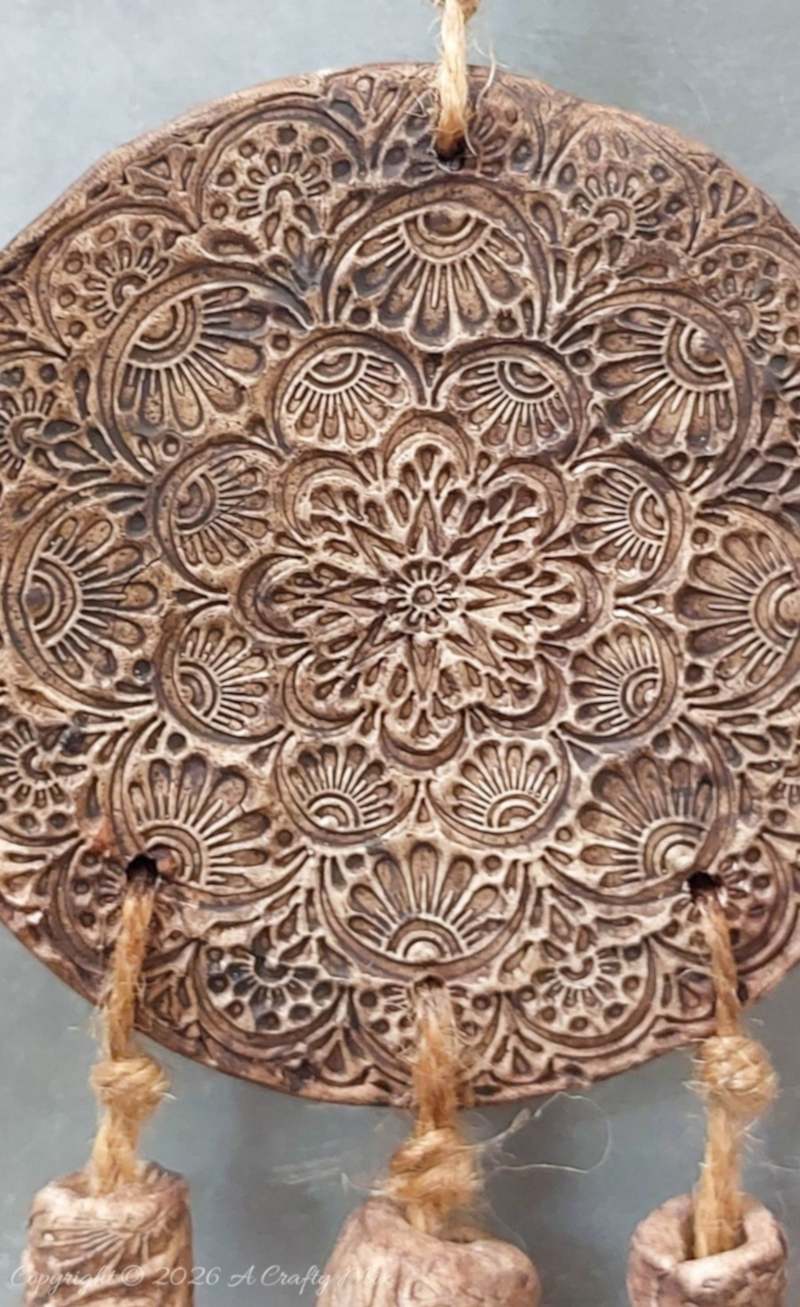 Close‑up of a round rustic clay medallion wall hanging with an intricate mandala imprint, aged brown finish, and jute twine strands threaded through holes at the bottom.