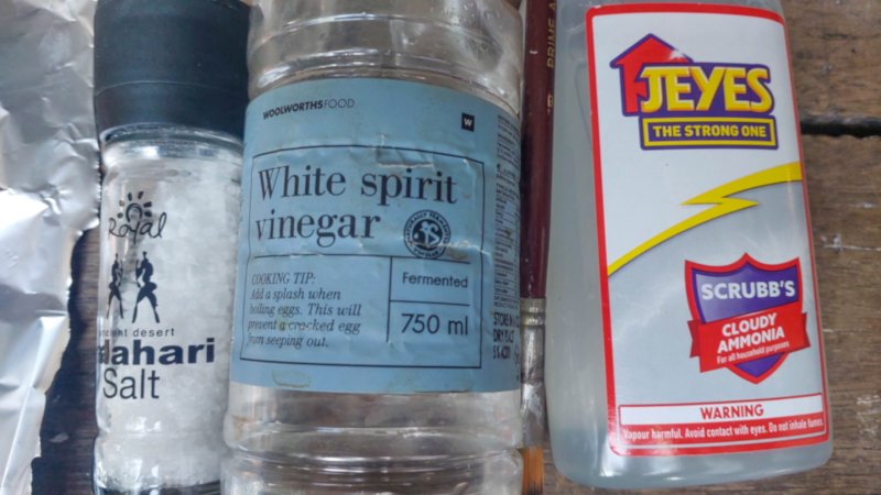 Three bottles containing coarse salt, vinegar, and ammonia lying on a wooden surface