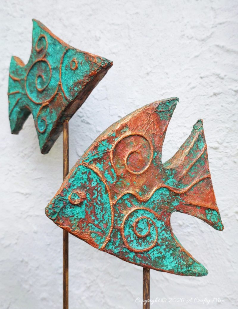 Two rustic patina fish on wooden sticks used as DIY coastal decor, crafted from layered cardboard and copper tape.