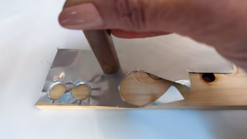 Hand pressing a short length of copper pipe into a sheet of aluminium on a wooden block to punch out small metal circles for soda can bird eyes.
