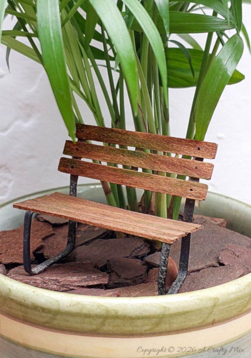 Mini wooden park bench with metal legs placed on bark chips inside a mug planter, with love palm (parlour palm) leaves rising up behind it.