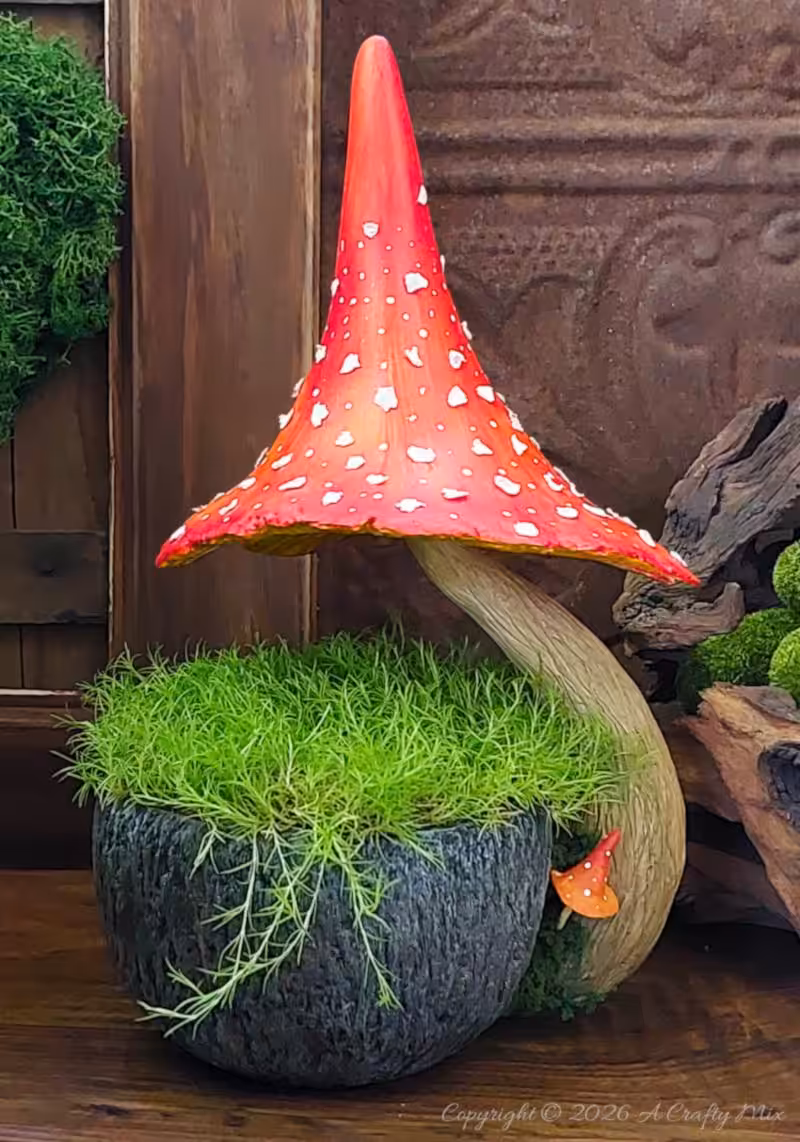 Whimsical fairy garden in a textured cup planter, topped with bright green moss and a large curved red mushroom with white spots, set against a rustic wooden backdrop.