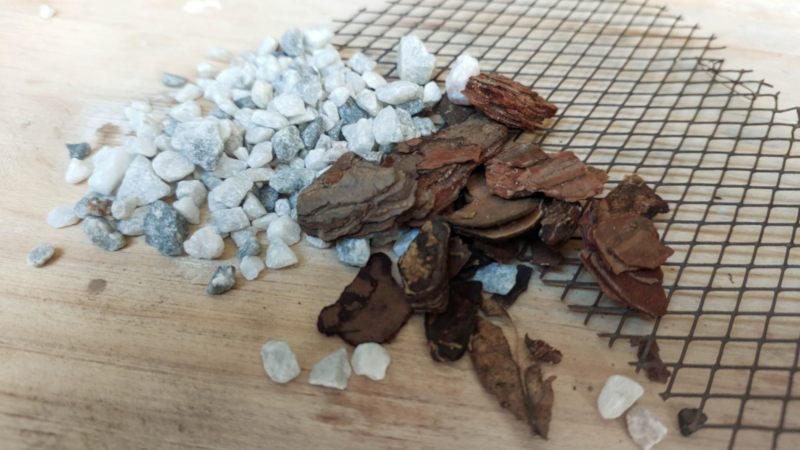 Small pile of white gravel and chunky bark pieces resting on top of a square of wire mesh, shown on a light wooden surface.