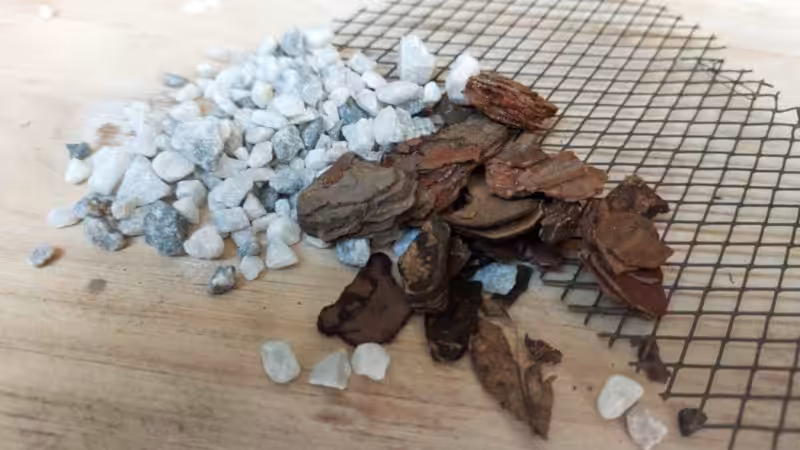 Small pile of white gravel and chunky bark pieces resting on top of a square of wire mesh, shown on a light wooden surface.