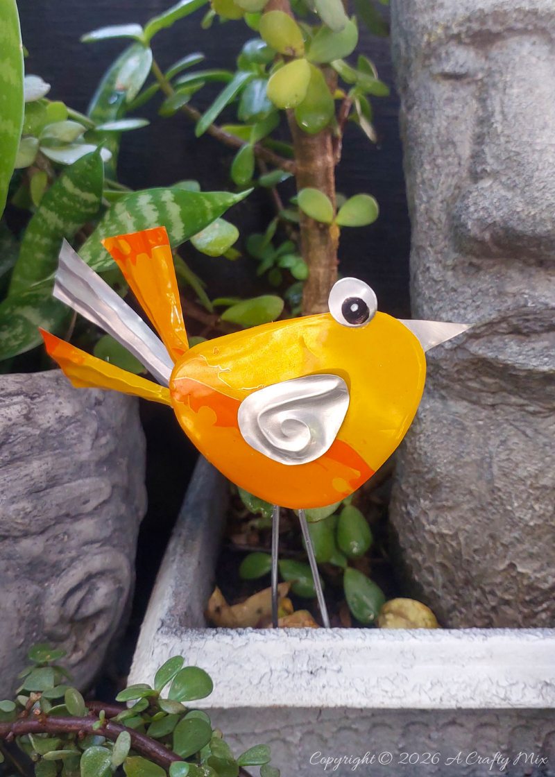 A bright orange, recycled Soda Can Bird on wire legs decorating a potted plant in a small garden.