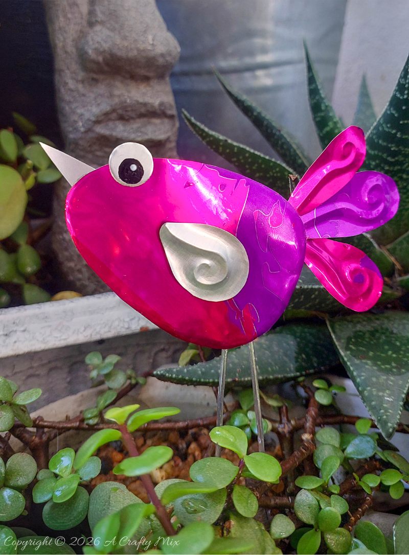 Close up of a purple and pink recycled Soda Can Bird made from an aluminium drink can with large eyes and embossed metal wings.
