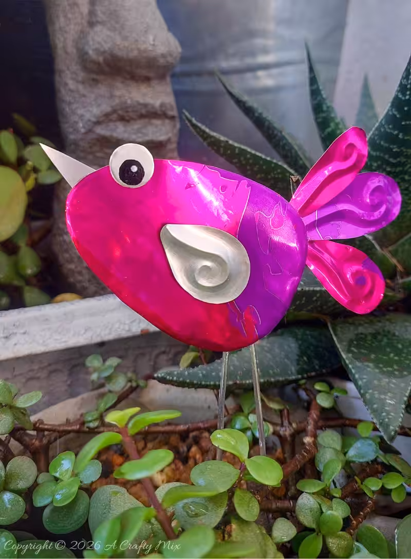 Close up of a purple and pink recycled Soda Can Bird made from an aluminium drink can with large eyes and embossed metal wings.