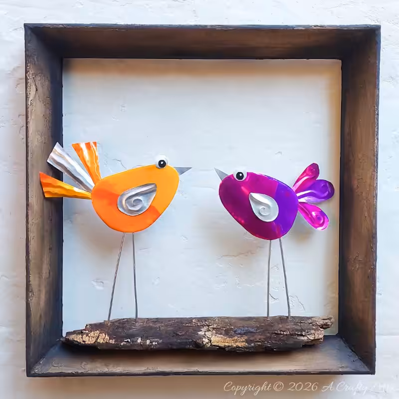Two colourful DIY Soda Can Birds made from recycled aluminium cans, standing on a piece of wood inside a rustic shadow‑box frame and displayed as wall art.