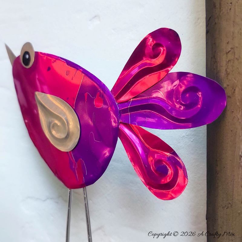 Close up of a purple and pink recycled Soda Can Bird made from an aluminium drink can with large eyes and embossed metal tail feathers and wings.