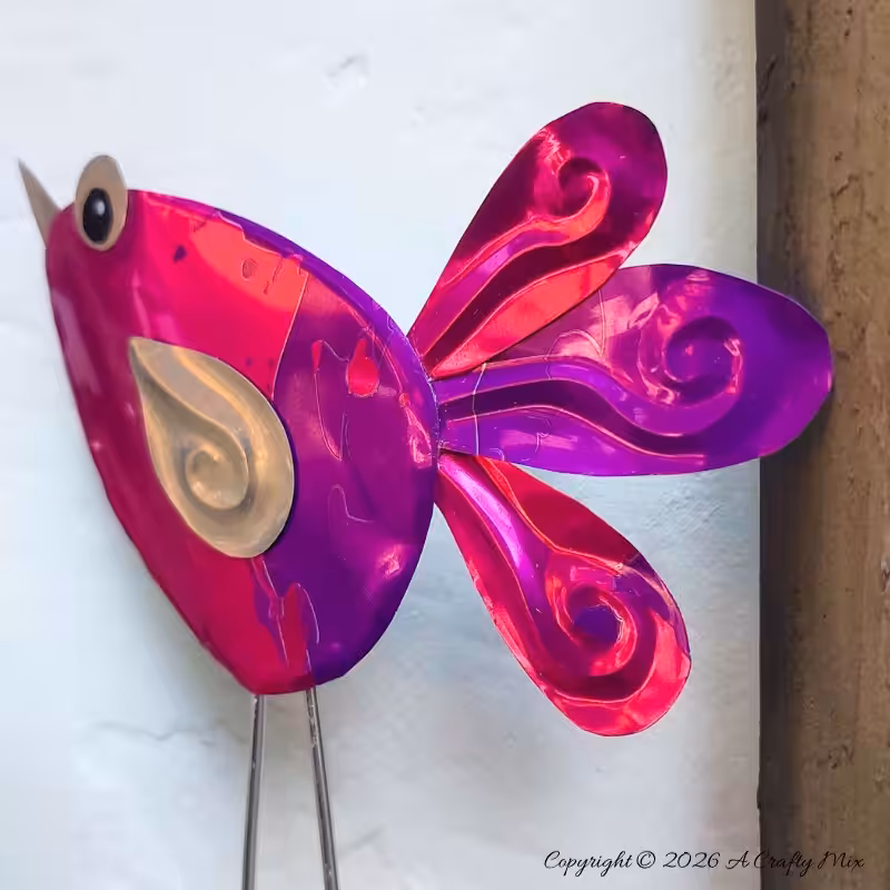 Close up of a purple and pink recycled Soda Can Bird made from an aluminium drink can with large eyes and embossed metal tail feathers and wings.
