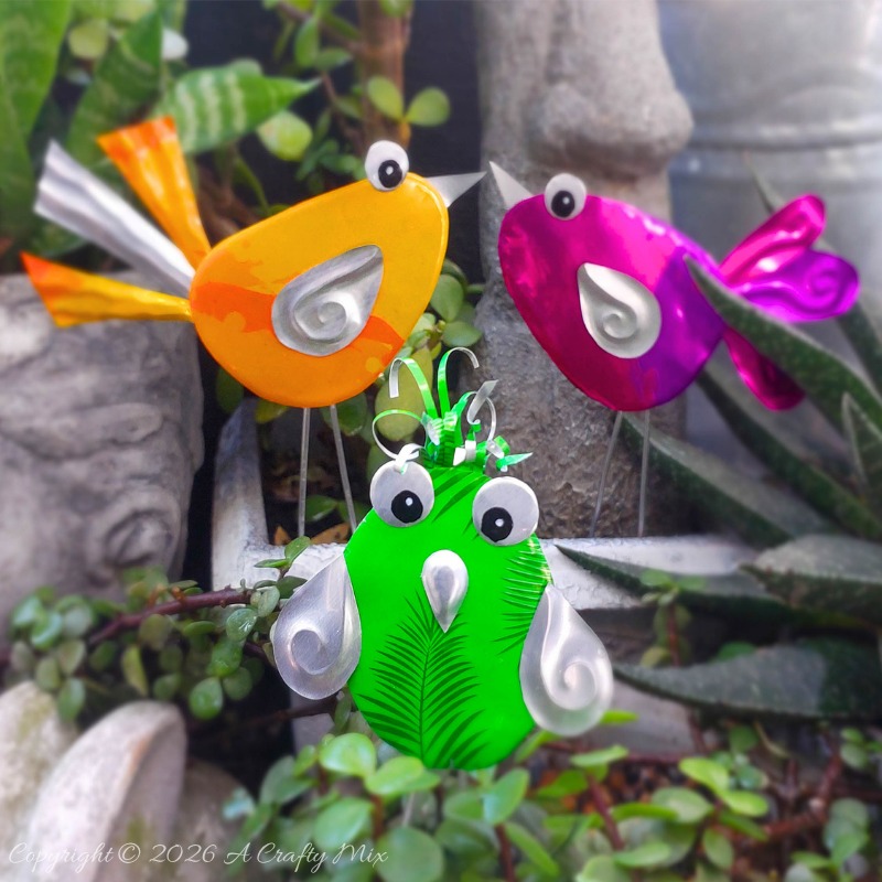 Orange, pink and green recycled Soda Can Birds on wire legs decorating potted plants in a small garden.