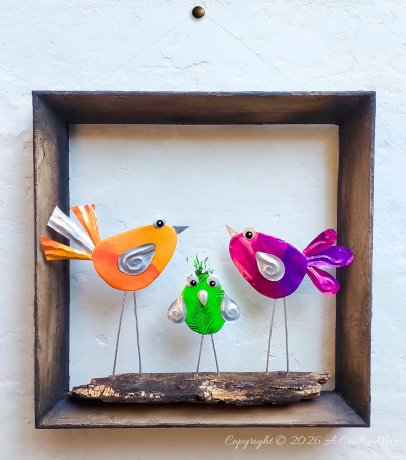 Three colourful DIY Soda Can Birds made from recycled aluminium cans, standing on a piece of wood inside a rustic shadow‑box frame and displayed as wall art.
