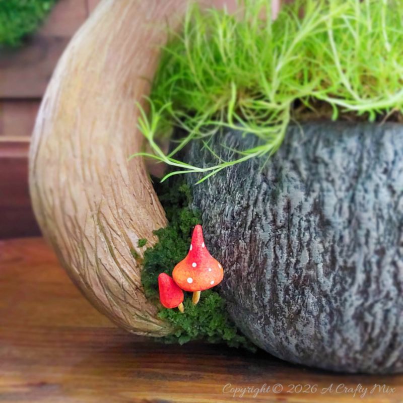 Close‑up of a fantasy teacup planter with bright green moss spilling over the rim and two tiny red mushrooms with white spots nestled at the base of a textured, tree‑like handle.