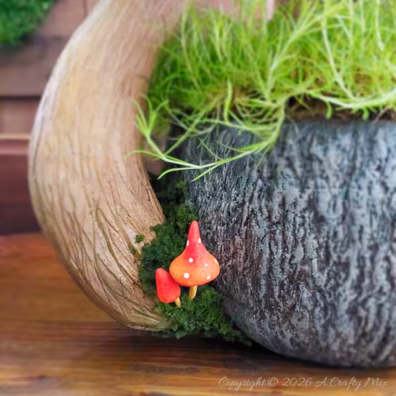 Close‑up of a fantasy teacup planter with bright green moss spilling over the rim and two tiny red mushrooms with white spots nestled at the base of a textured, tree‑like handle.