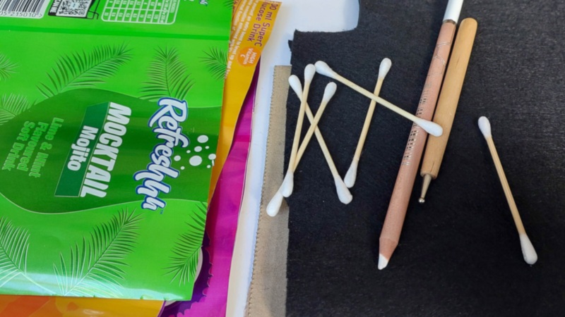 Supplies laid out for a soda can bird craft, including flattened colourful soda cans, cotton buds, embossing tools and pencils on a black felt work surface.