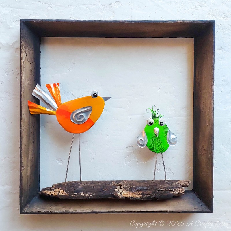 Two colourful DIY Soda Can Birds made from recycled aluminium cans, standing on a piece of wood inside a rustic shadow‑box frame and displayed as wall art.