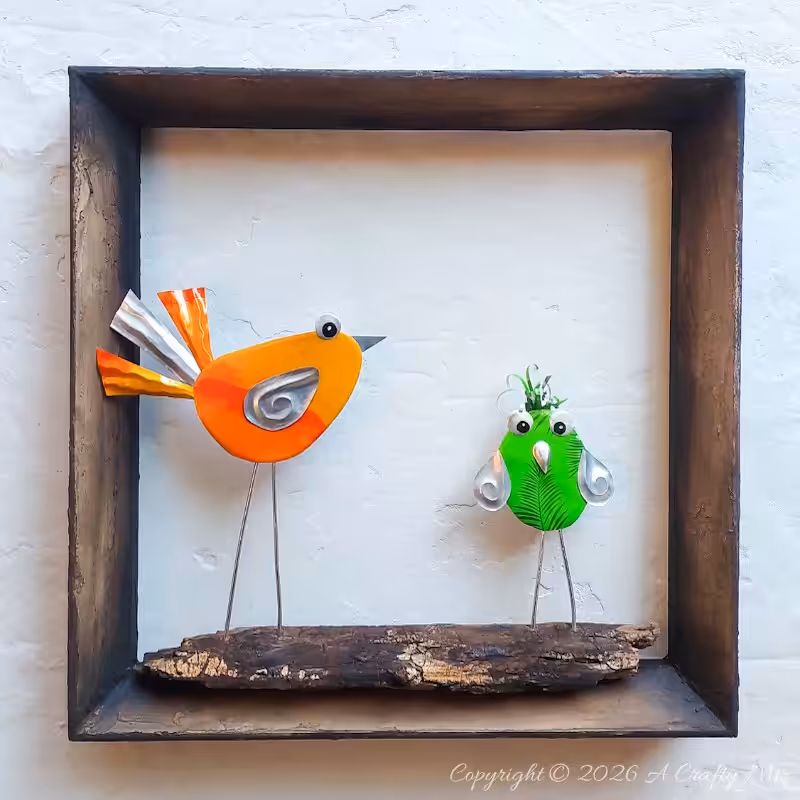 Two colourful DIY Soda Can Birds made from recycled aluminium cans, standing on a piece of wood inside a rustic shadow‑box frame and displayed as wall art.