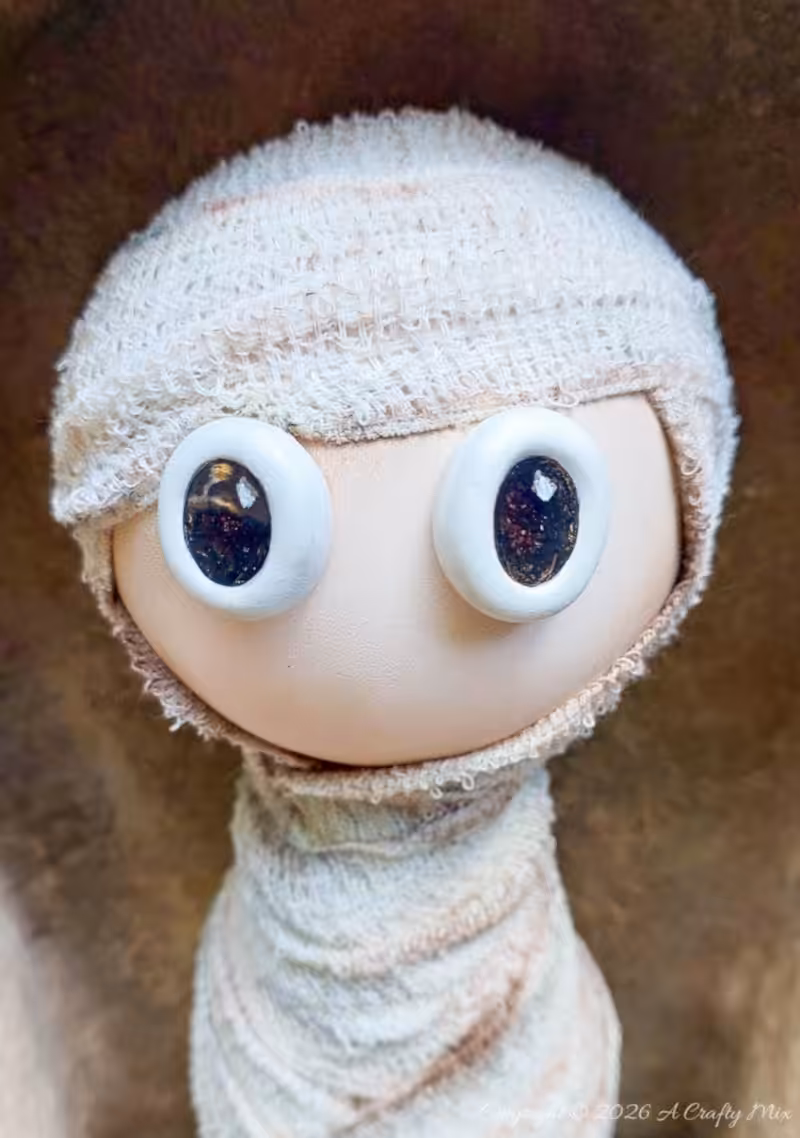 Close-up of a handmade Halloween mummy with large glossy black-and-white eyes, a smooth beige face, and aged white bandages wrapped around its head and body.