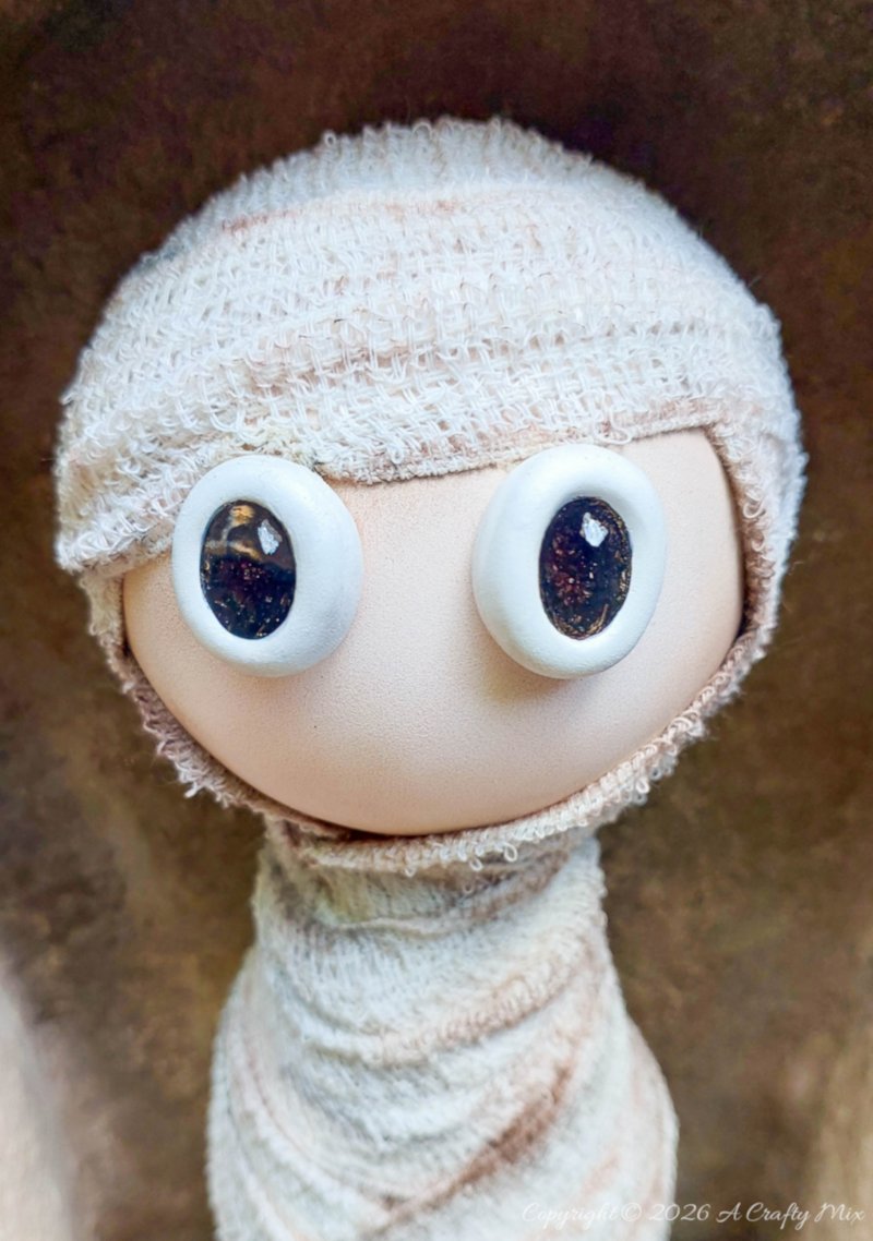 Close-up of a handmade Halloween mummy with large glossy black-and-white eyes, a smooth beige face, and aged white bandages wrapped around its head and body.