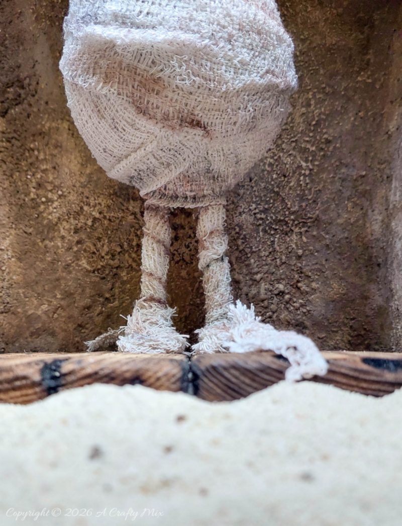 Close-up of an adorable handmade mummy’s thin legs and frayed bandage feet standing on a faux wooden floor inside a textured stone sarcophagus.