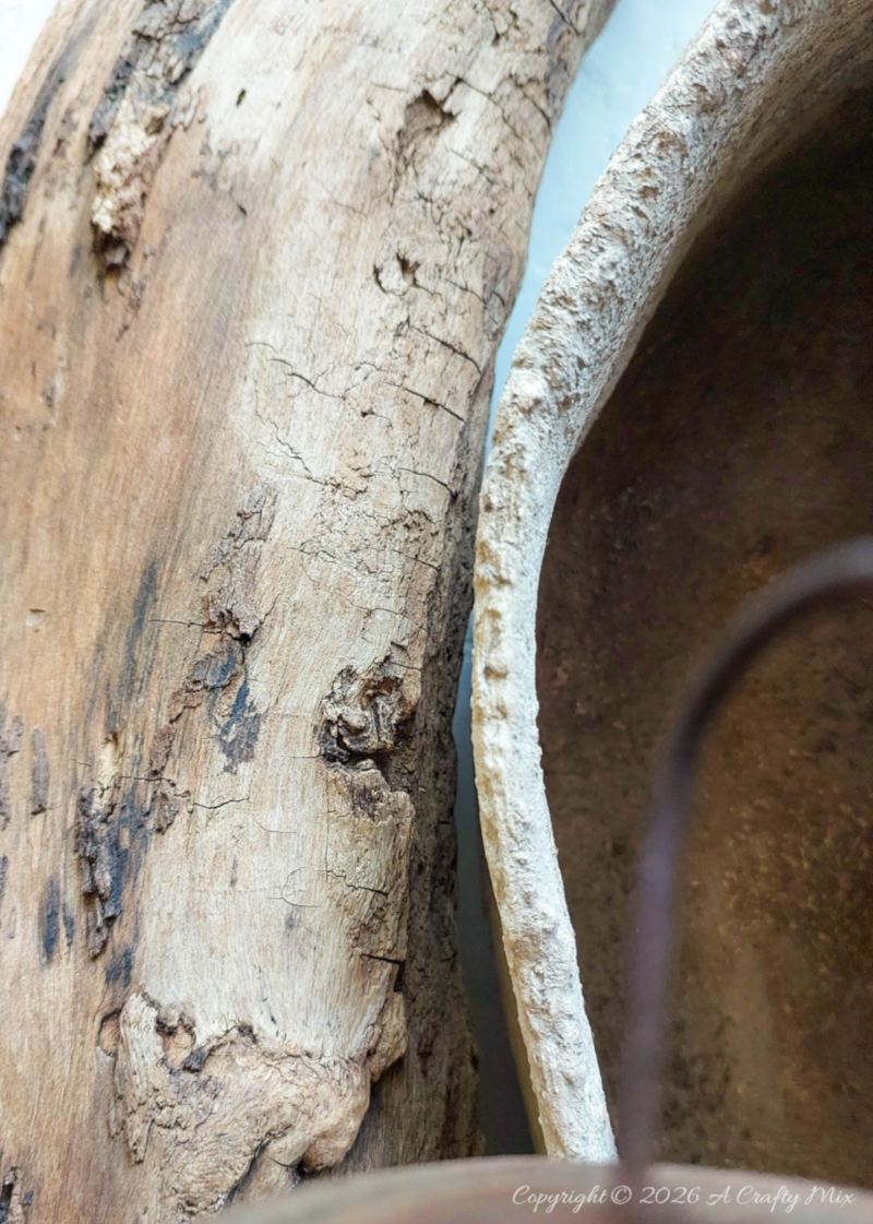 Close-up of the rough, pitted edge of a handmade faux stone cardboard sarcophagus beside weathered driftwood.