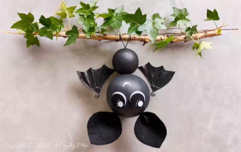 Small black DIY bat hanging upside down from an ivy-covered branch, with shiny crinkled plastic bag bat wings and oversized white-and-black eyes