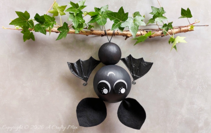 Small black DIY bat hanging upside down from an ivy-covered branch, with shiny crinkled plastic bag bat wings and oversized white-and-black eyes