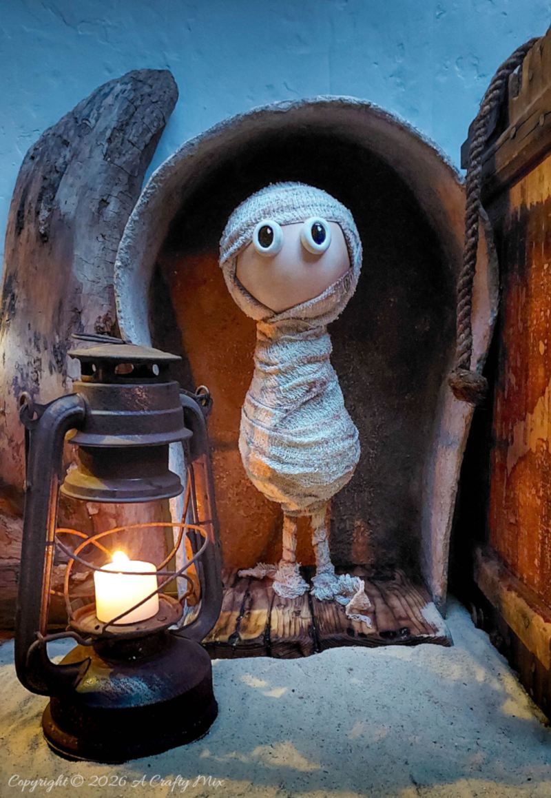 Handmade white bandage mummy standing in a faux stone sarcophagus beside a rusty lantern with a glowing candle, photographed in low evening light.