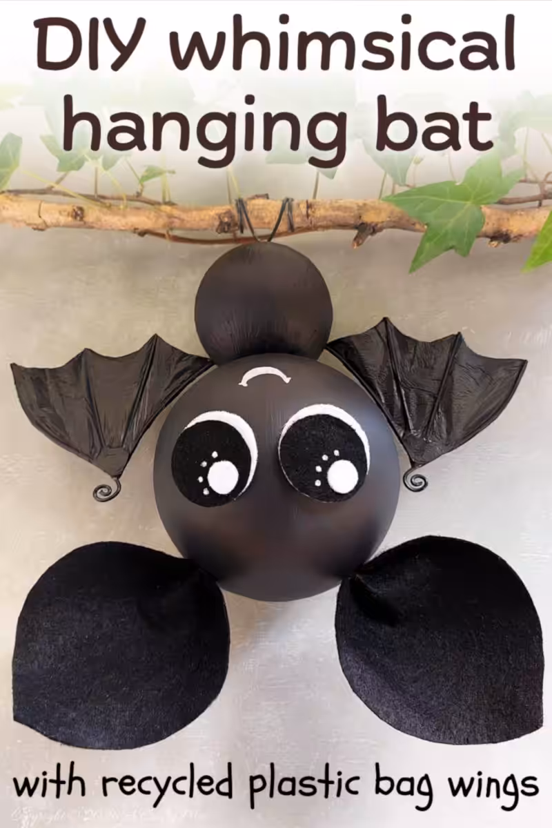 Pin graphic of a cute black hanging bat with large eyes, recycled plastic bag bat wings, and black wire feet, hanging from an ivy-covered branch.