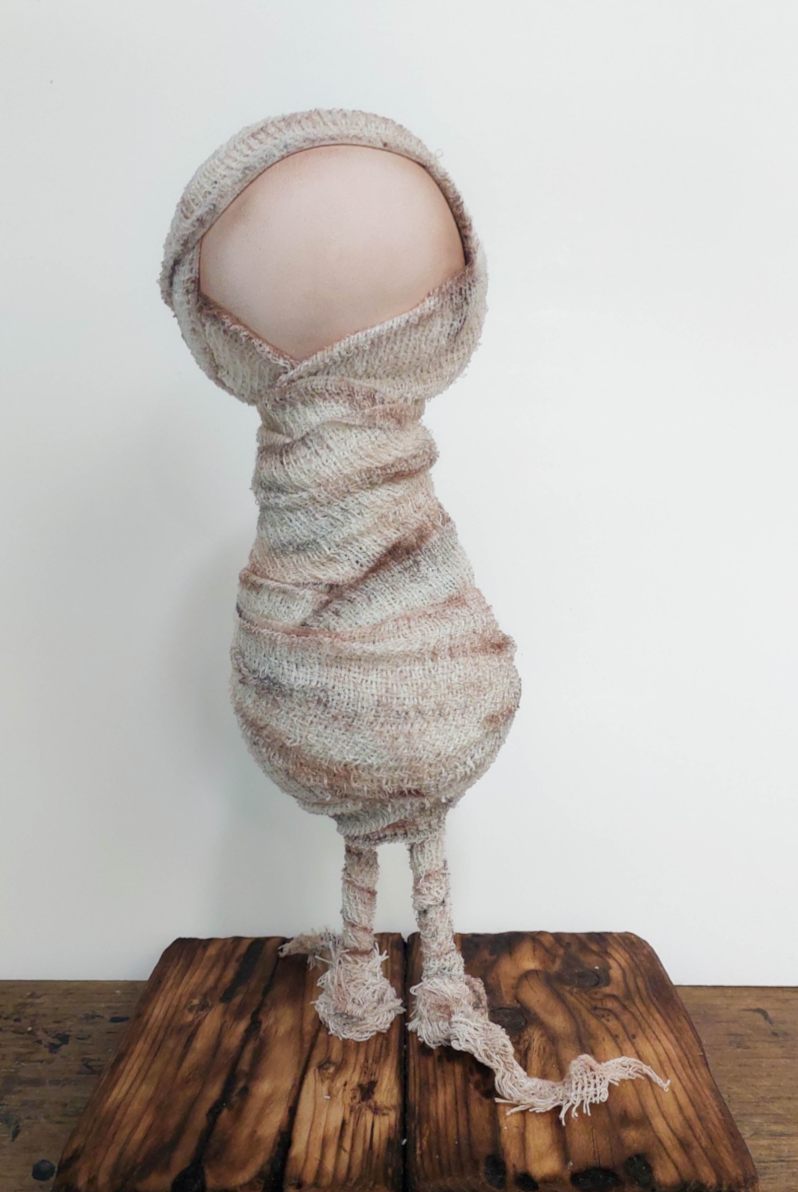 Eyeless handmade Halloween mummy wrapped in aged off-white bandages, standing on a dark faux wooden floor with long skinny legs and frayed fabric feet.