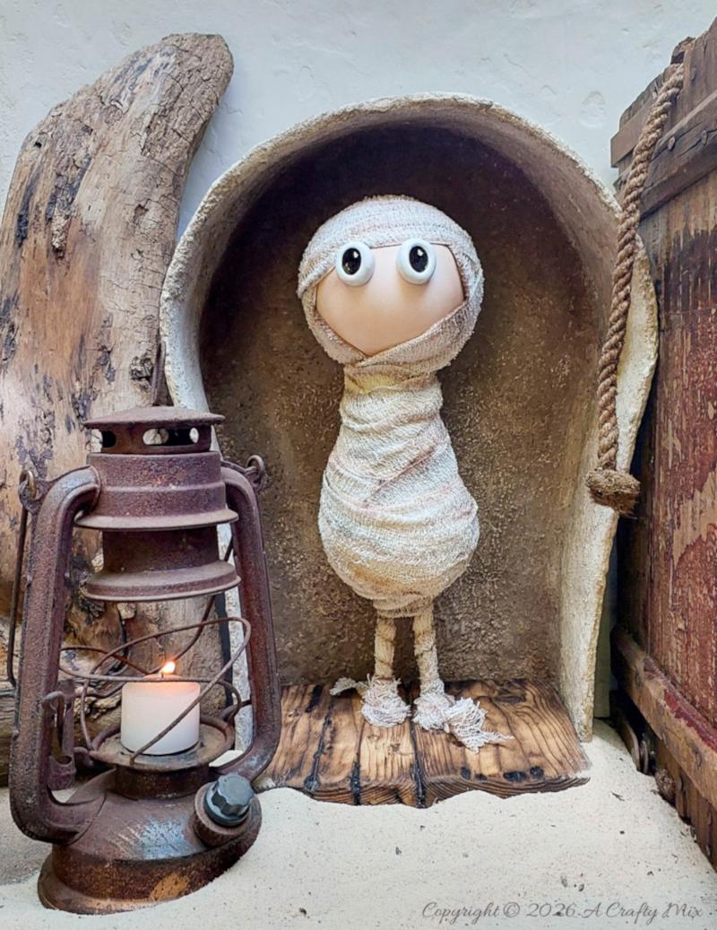 Handmade bandage mummy with large eyes standing inside a faux stone sarcophagus beside a rusty lantern, driftwood, and an old wooden crate.