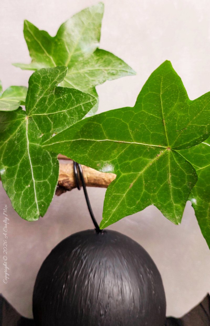 Whimsical black DIY hanging bat with round eyes, plastic bag wings, and felt feet suspended from an ivy-covered branch.