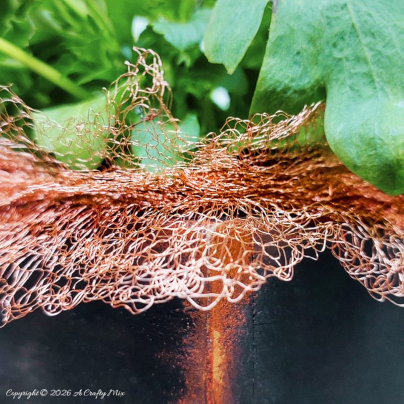 Detail shot of copper mesh that will help keep snails away, draped over the rim of a dark wooden planter, with bright green foliage above
