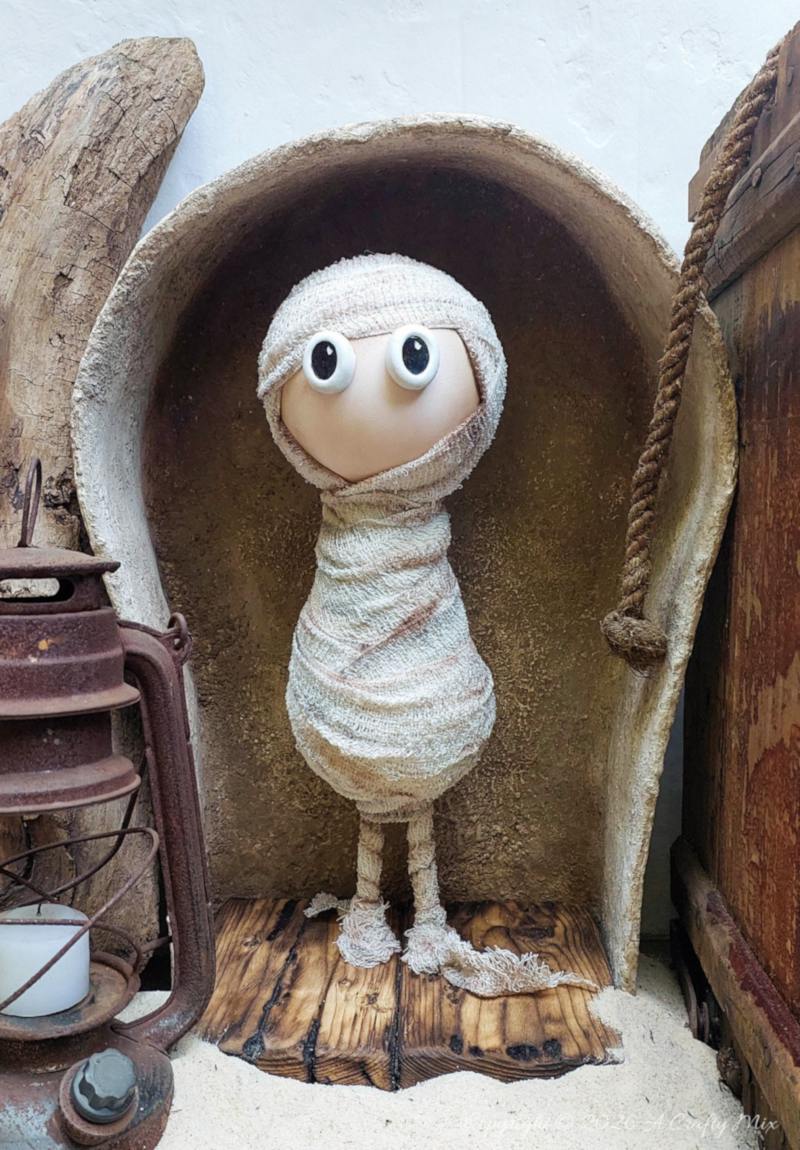 Full view of a cute handmade mummy with oversized eyes standing in a large, textured faux stone sarcophagus on a wooden floor, beside a rusty lantern.