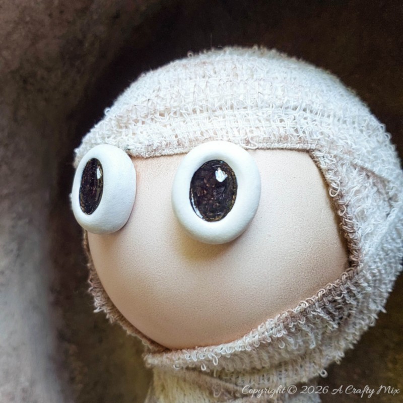 Close-up side view of a handmade plastic bottle mummy with a smooth beige face, large shiny black-and-white eyes, and frayed off-white bandages wrapped around its head.