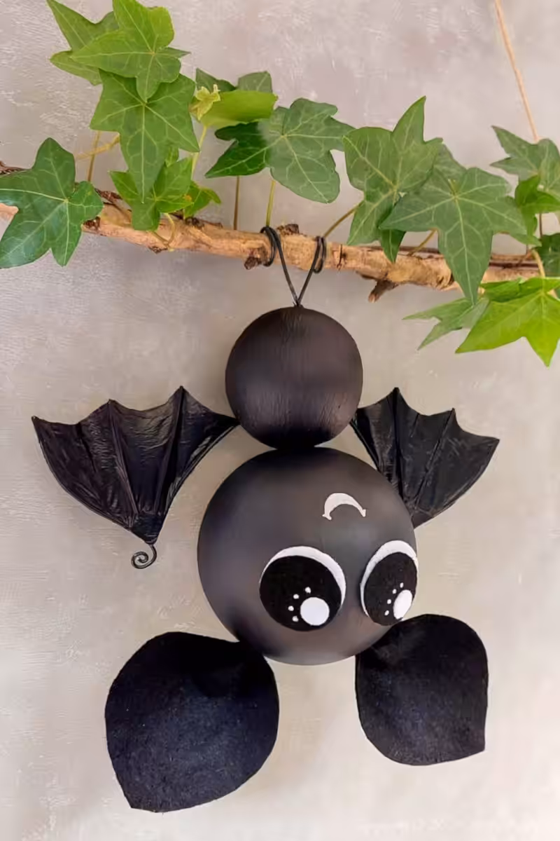 Side view of a black hanging bat with oversized eyes, crinkled plastic bag bat wings, and black felt feet beneath an ivy-covered branch.