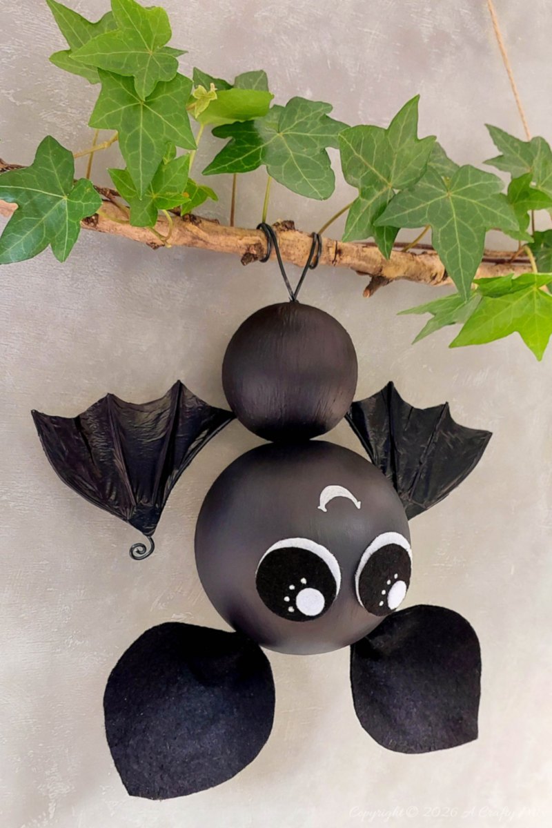Side view of a black hanging bat with oversized eyes, crinkled plastic bag bat wings, and black felt feet beneath an ivy-covered branch.