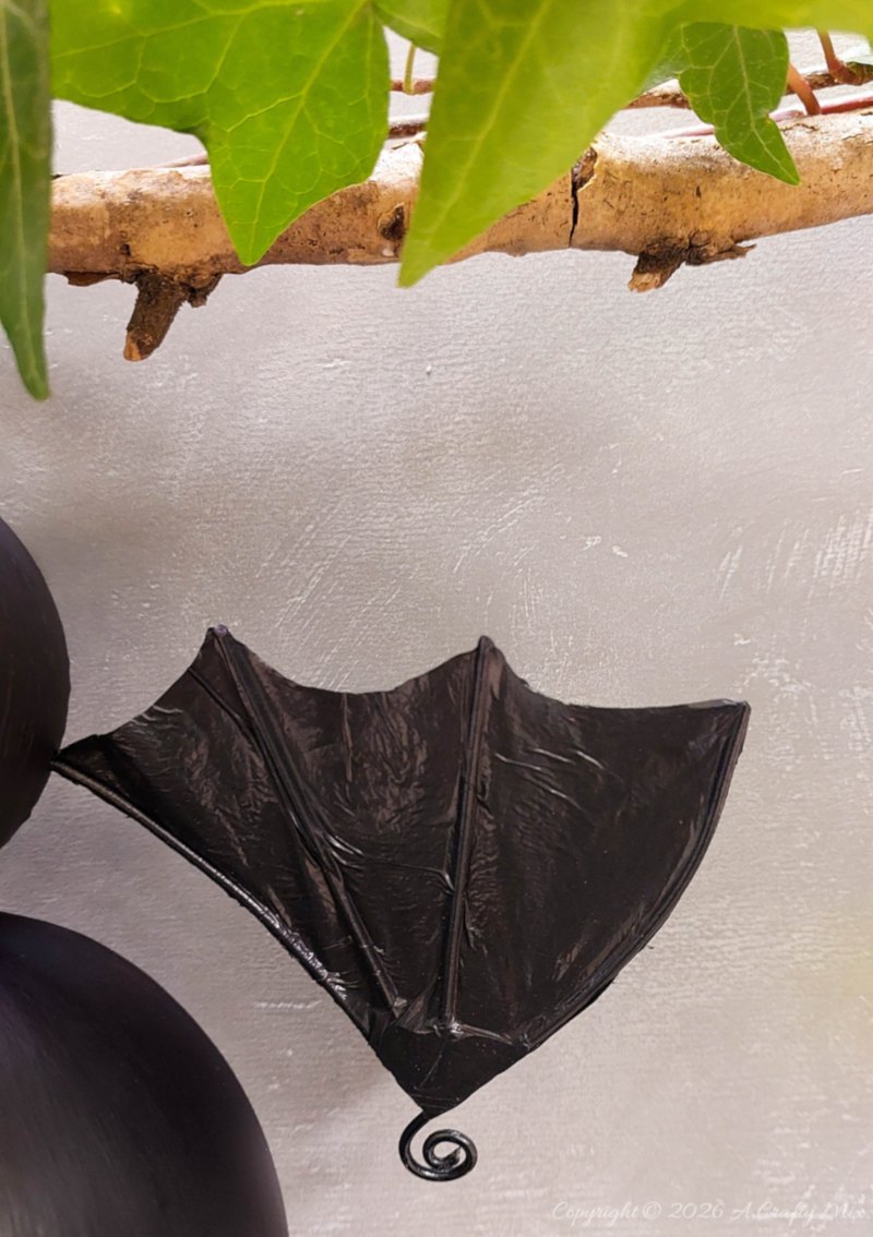 Black DIY hanging bat with large white-and-black eyes and textured plastic bag wings hanging from a branch with green ivy.